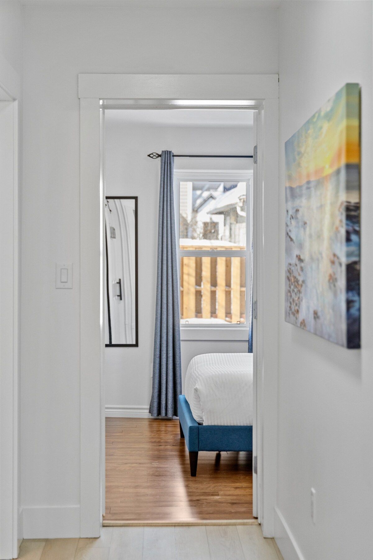 Ein schmaler Flur führt zu einem Schlafzimmer mit Bett, Fenster mit Blick auf eine Holzterrasse und einem Bild an der Wand.