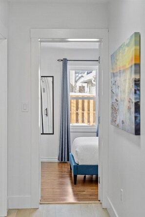 Ein schmaler Flur führt zu einem Schlafzimmer mit Bett, Fenster mit Blick auf eine Holzterrasse und einem Bild an der Wand.