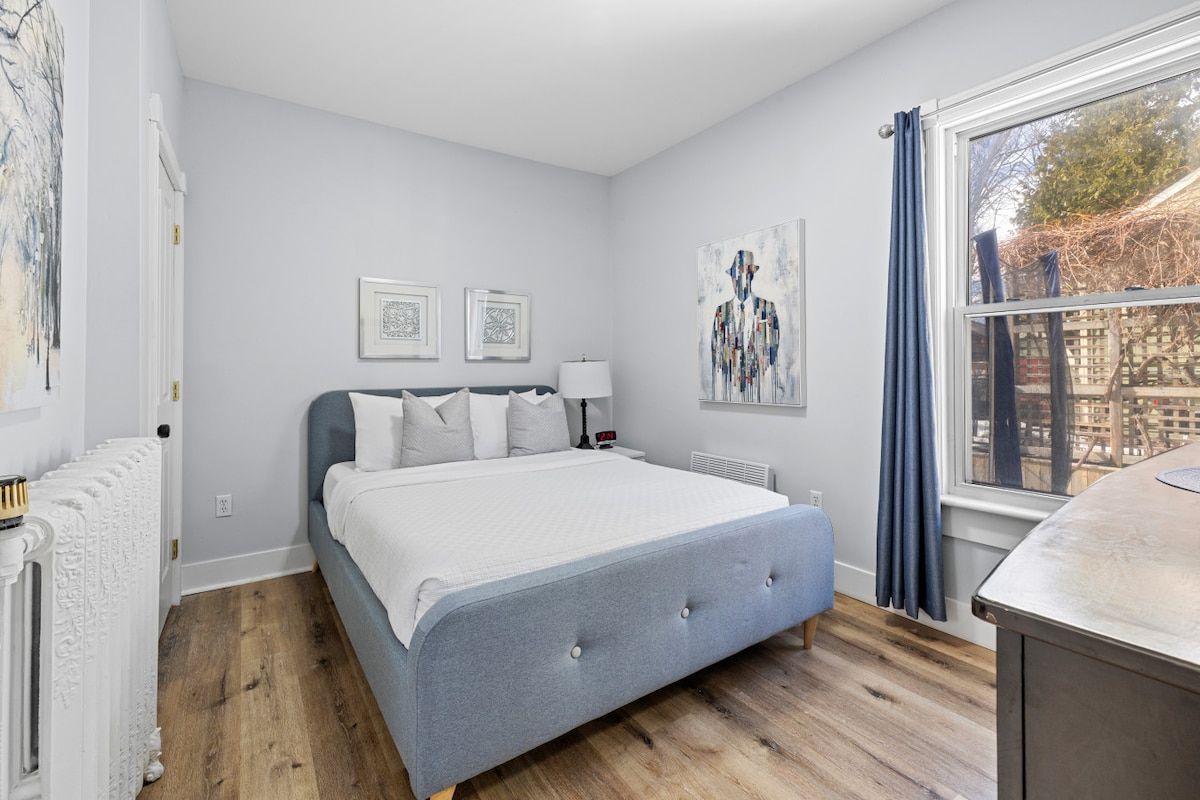 Een slaapkamer met een bed, een radiator, een raam met gordijnen en kunstwerken aan de wanden.