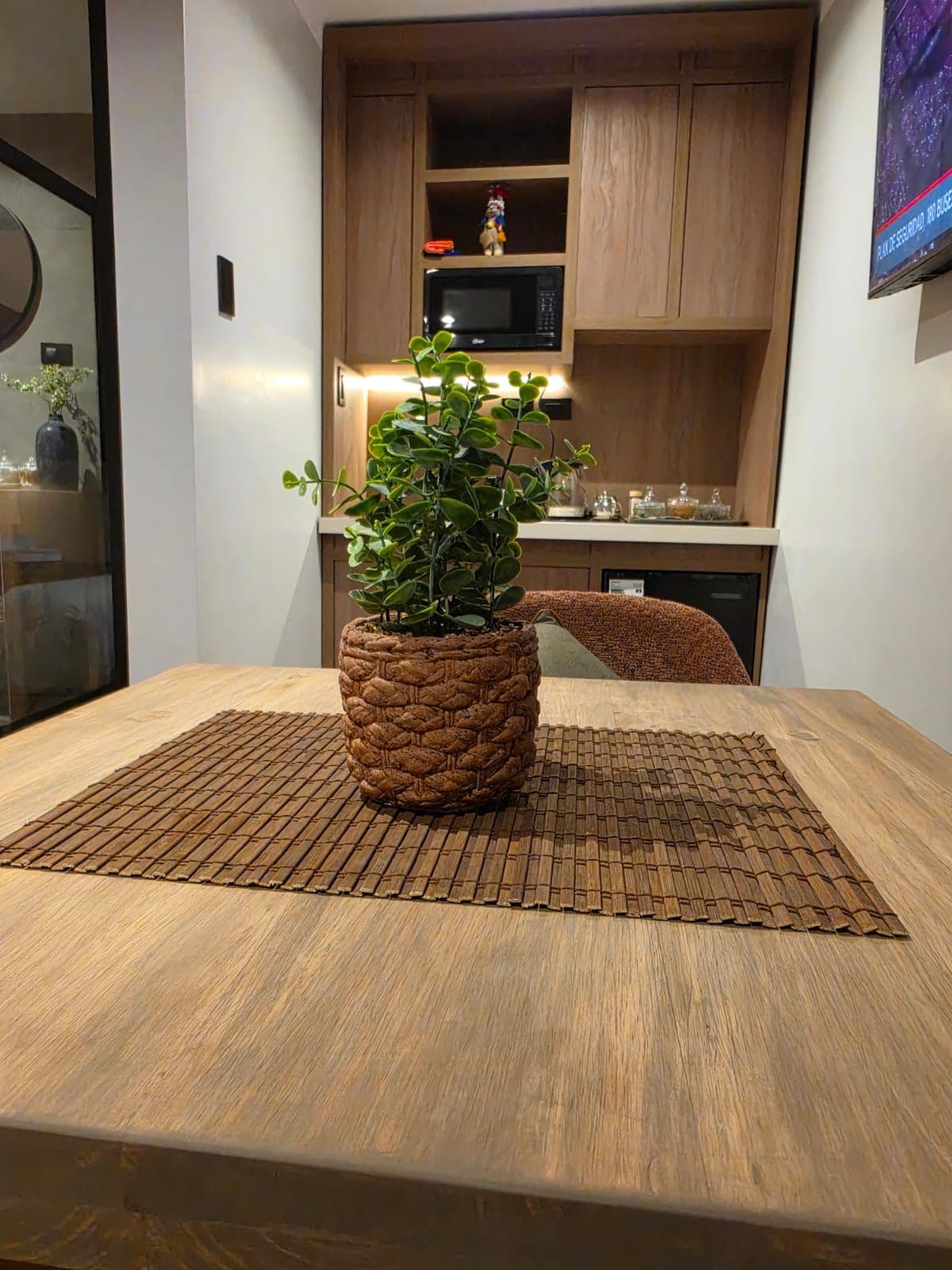 Une table à manger avec un set de table en fibres tressées, une plante en pot et un meuble en bois en arrière-plan.