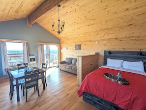 Ein Zimmer mit Holzbalkendecke, einem Bett mit roter Bettwäsche, einem Esstisch mit Stühlen und Blick durch ein großes Fenster.