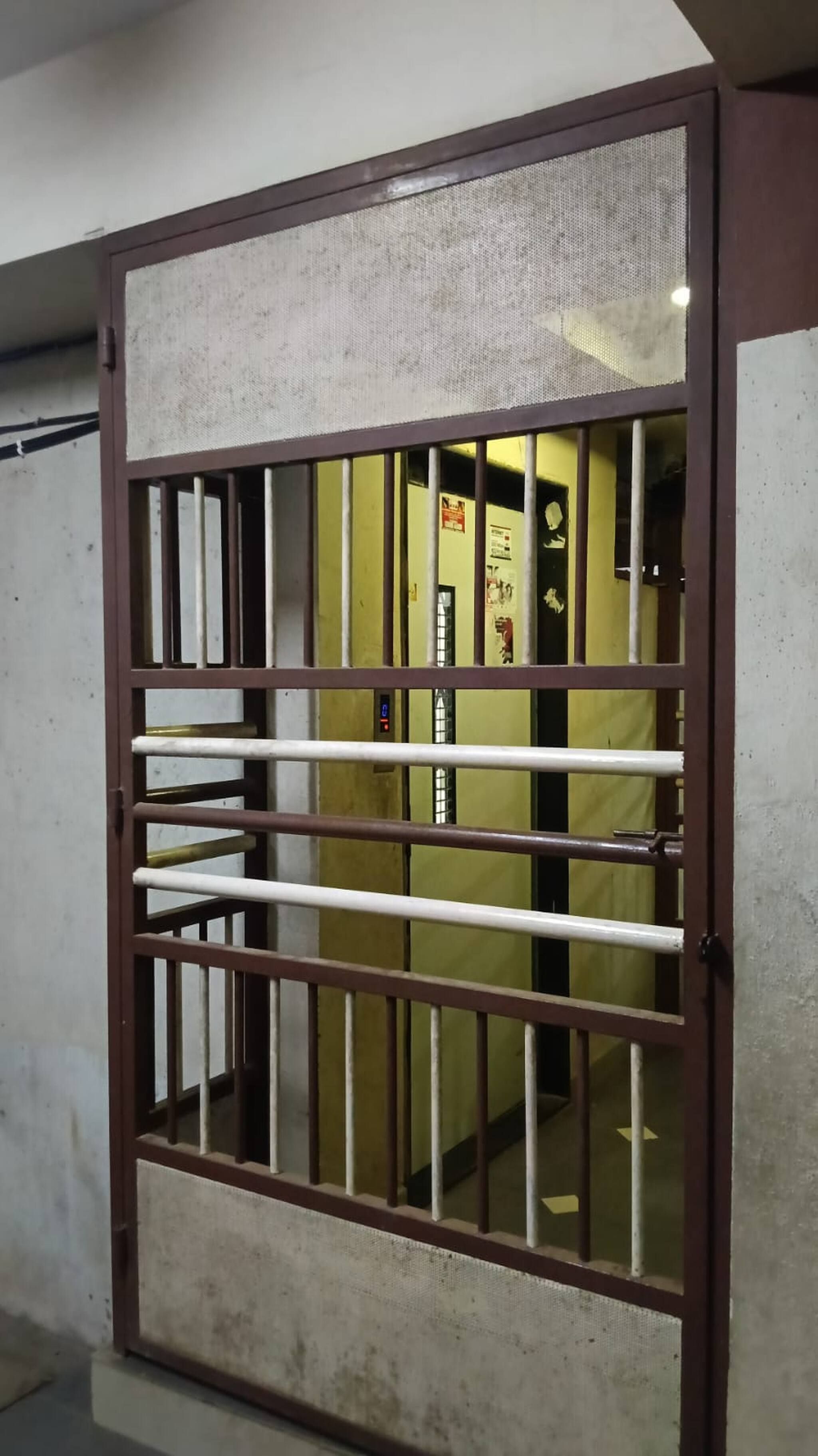 Una ventana con rejas de metal, un panel de control de ascensor visible y una pared con un cartel.