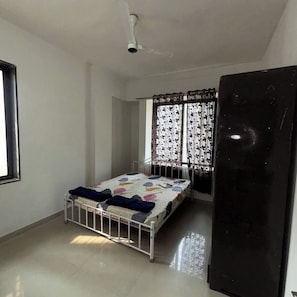 A room with a bed, a ceiling fan, a window with patterned curtains, and a black refrigerator. - Comfortable 3 BHK Homestay (Nashik)