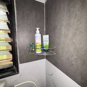 A bathroom corner with a grey tiled wall, a metal shelf holding toiletries, and a window with blinds partially visible.