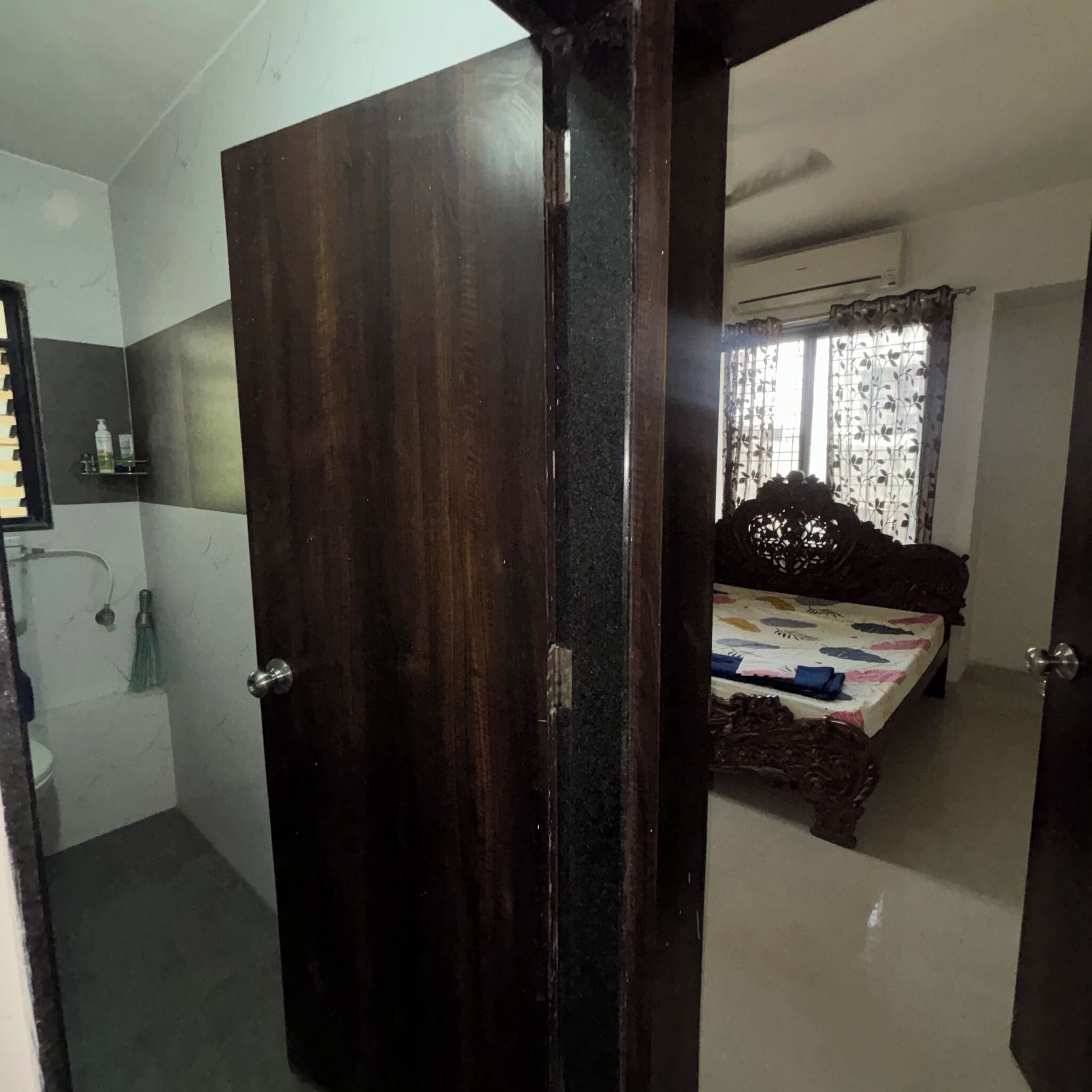 Un dormitorio con puerta de madera, cama con colcha floral, ventana con cortinas y una pequeña repisa con artículos de aseo.
