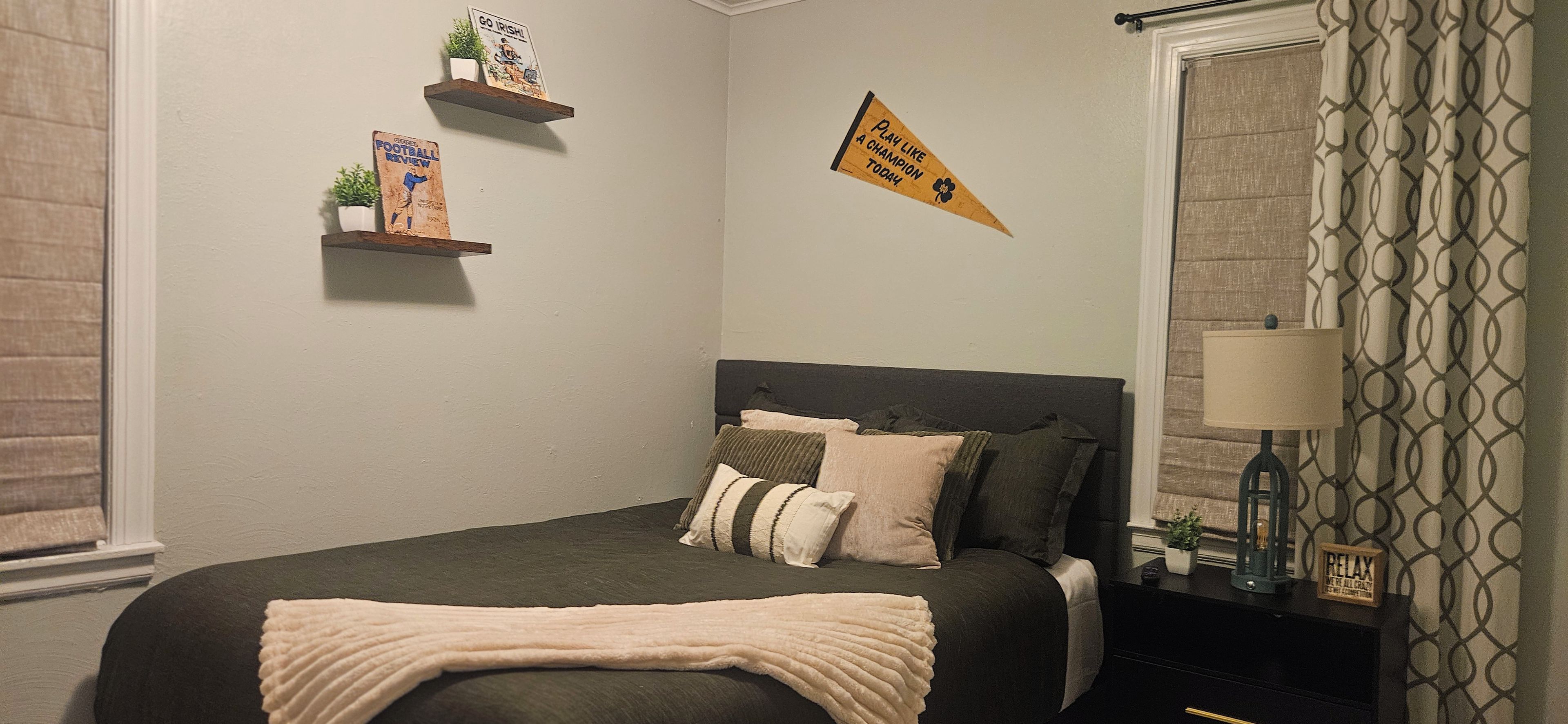 A bedroom with a bed, pillows, a bedside table with a lamp, a wall-mounted shelf with books, and a window with curtains.