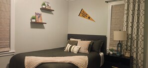 Ein Schlafzimmer mit einem Bett, Kissen, einem Nachttisch mit Lampe, einem an der Wand befestigten Regal mit Büchern und einem Fenster mit Vorhängen.