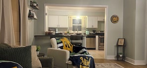 Ein Wohnzimmer mit einer Couch, einem Couchtisch mit einer Pflanze und Blick in eine Küche mit weißen Schränken und einem Essbereich.