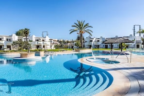 A swimming pool with a circular design, surrounded by a paved area with palm trees and a white building with a tiled roof in the background. - New Listing (Albufeira)