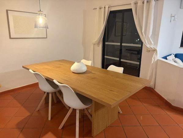 A dining area with a wooden table, white chairs, a white vase, a pendant light, and a sliding glass door. - New Listing (Albufeira)