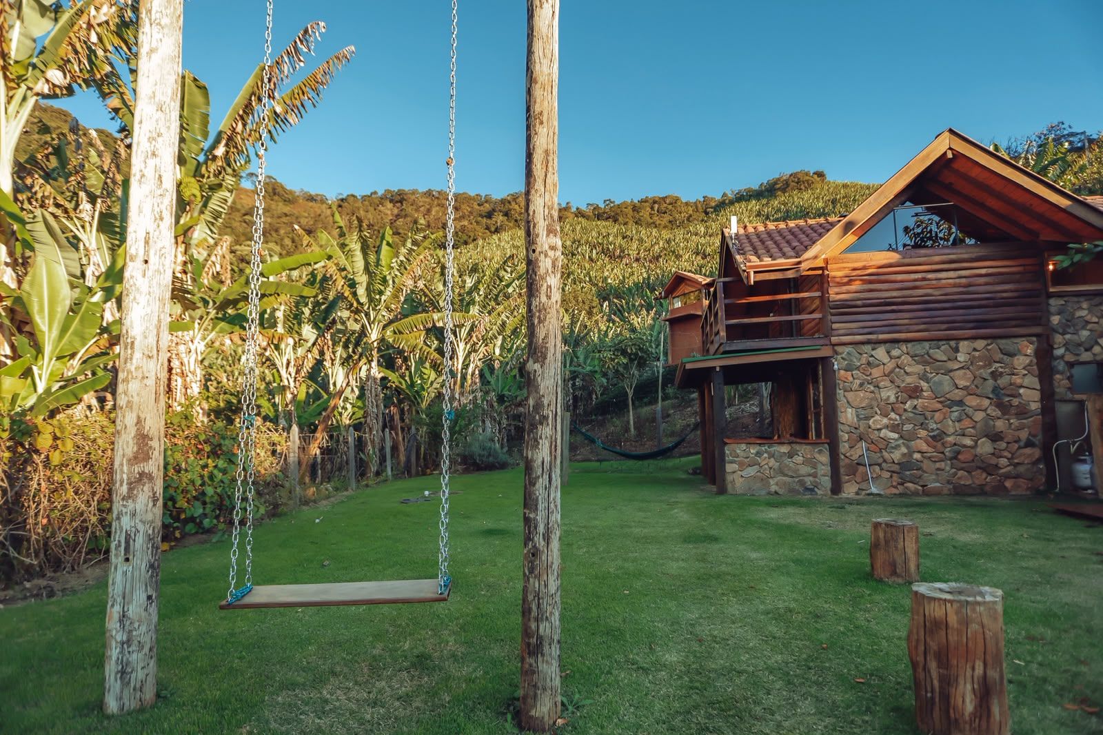 Une maison de campagne au charme rustique, avec des fondations en pierre, une terrasse en bois et un ensemble de balançoires dans un jardin luxuriant.