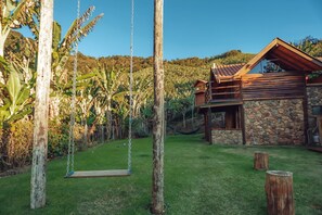 A rustic house with a stone foundation, a wooden deck, and a swing set in a lush garden. - Cabanas com Hidromassagem- Jeito da Roça (São Bento do Sapucaí)
