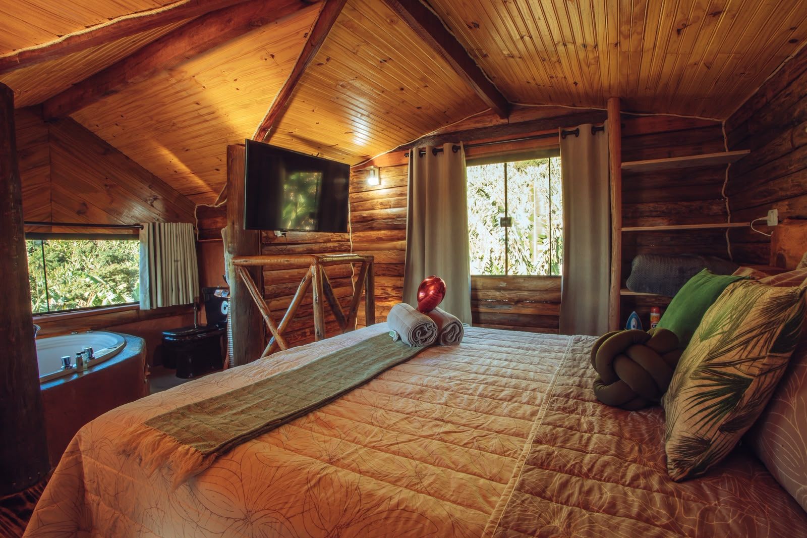 Ein Zimmer einer Holzhütte mit einem Bett, einem Fernseher, einem Fenster mit Vorhängen und Blick auf Grünfläche vor dem Fenster.
