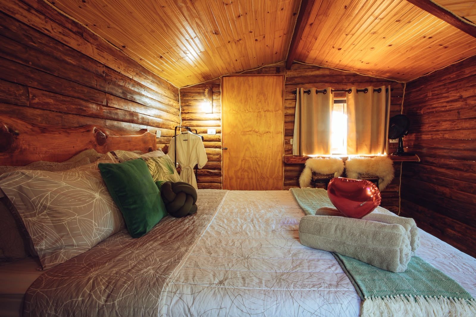 Une chambre confortable dans un chalet en rondins, avec un lit, des oreillers, un peignoir suspendu et une fenêtre avec des rideaux.