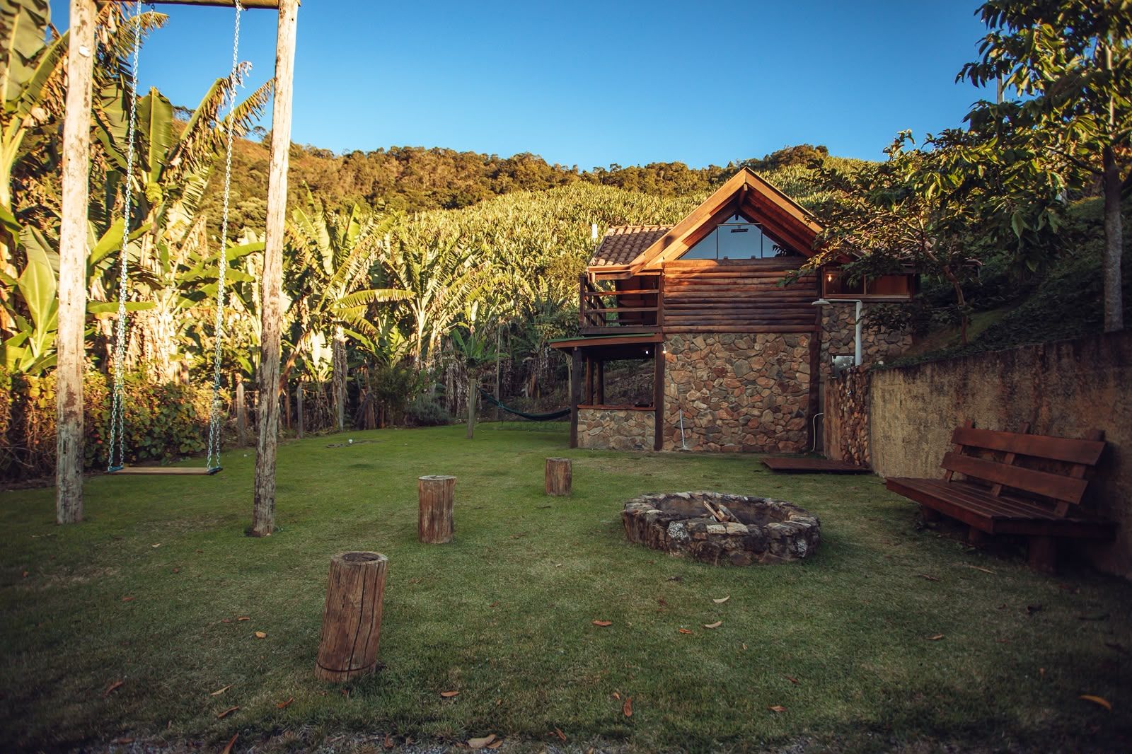 A rustic cabin with a stone exterior, a wooden deck, and a fire pit in a lush garden.