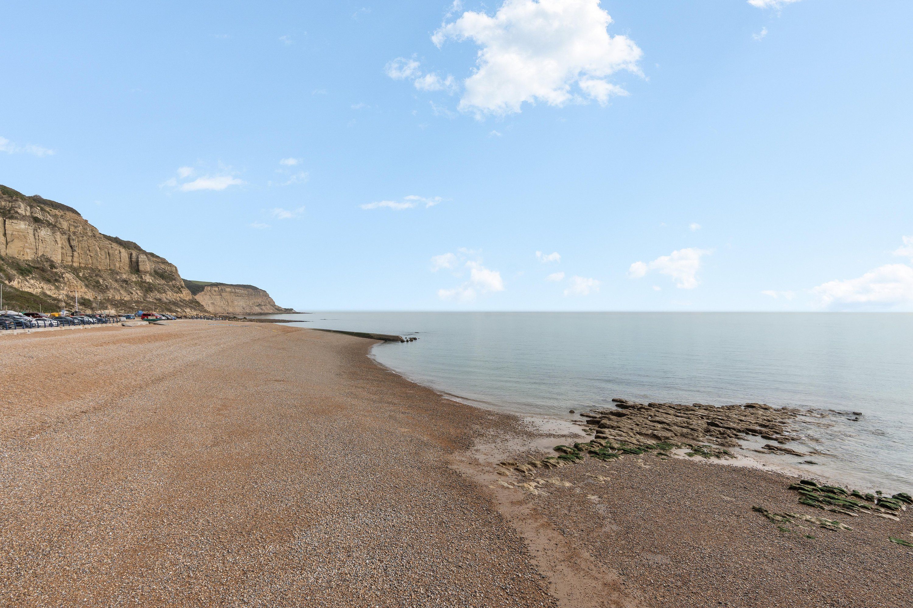 A pebble beach with a calm sea, a cliff, and a row of parked cars.