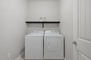 Ein Waschraum mit zwei weißen Whirlpool Waschmaschinen und Trocknern, einem schwarzen Regal darüber und einer weißen Tür rechts.