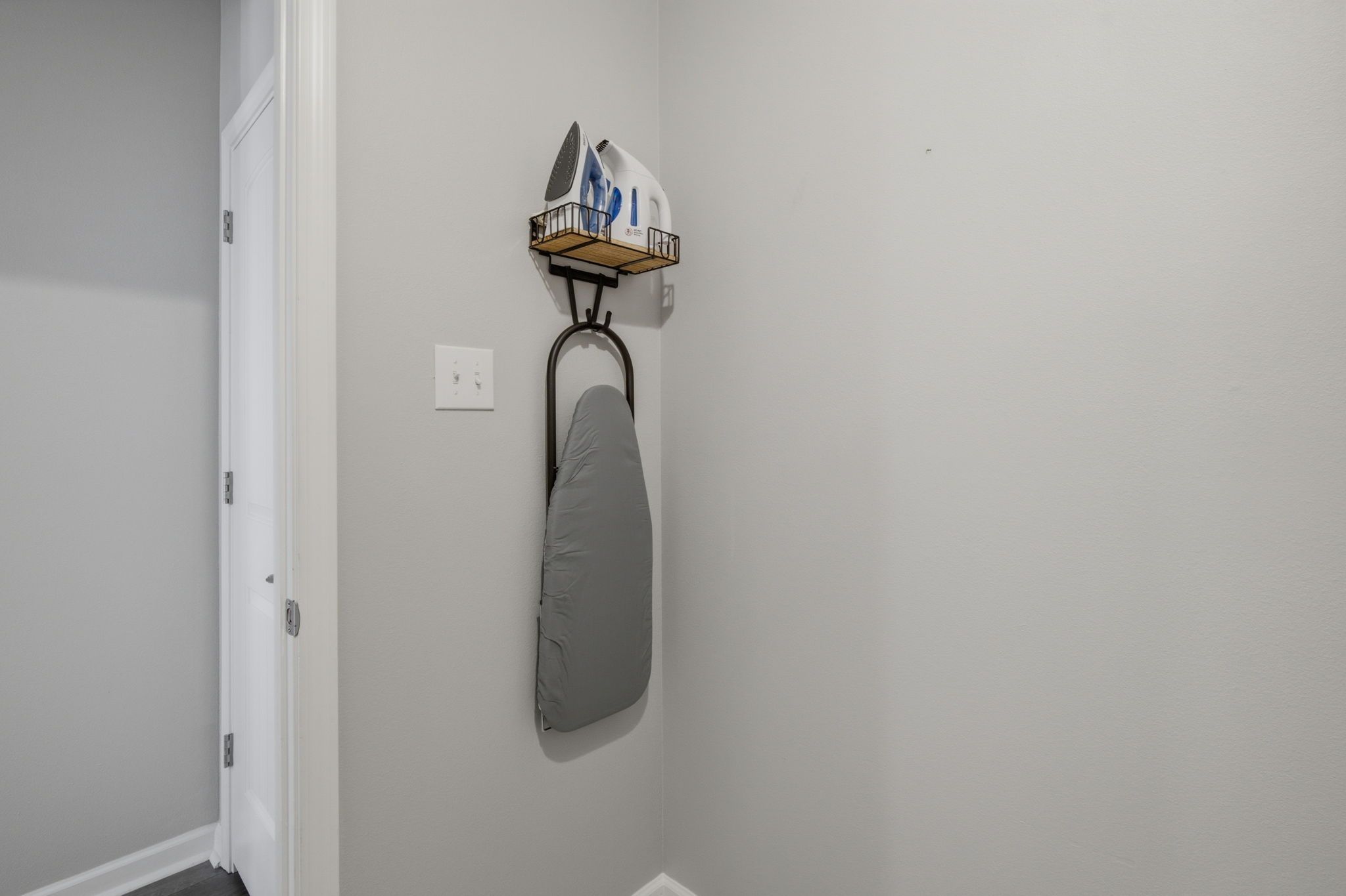 Een hoek van een kamer met een witte deur, een wandmontage strijkplank en een plankje met een waterkoker en andere spullen.