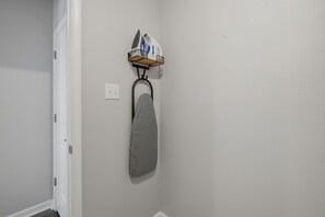 Een hoek van een kamer met een witte deur, een wandmontage strijkplank en een plankje met een waterkoker en andere spullen.