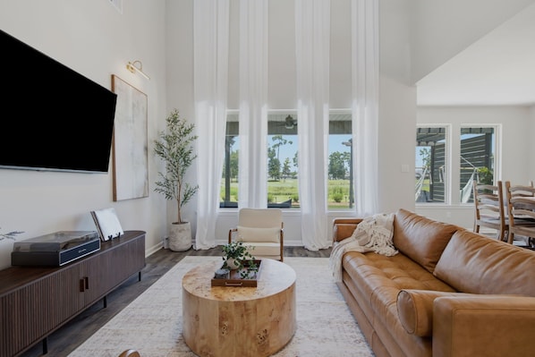 A modern living room with a leather sofa, a round wooden coffee table, a flat-screen TV, and a large window with curtains. - Golf Course Retreat | 2 Golf Carts + Theater Room (Westlake)