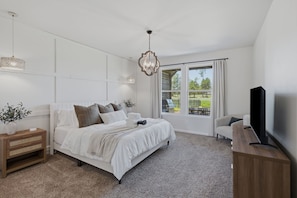 A bedroom with a large bed, a TV on a wooden stand, a window with curtains, and a plant on the nightstand. - Golf Course Retreat | 2 Golf Carts + Theater Room (Westlake)
