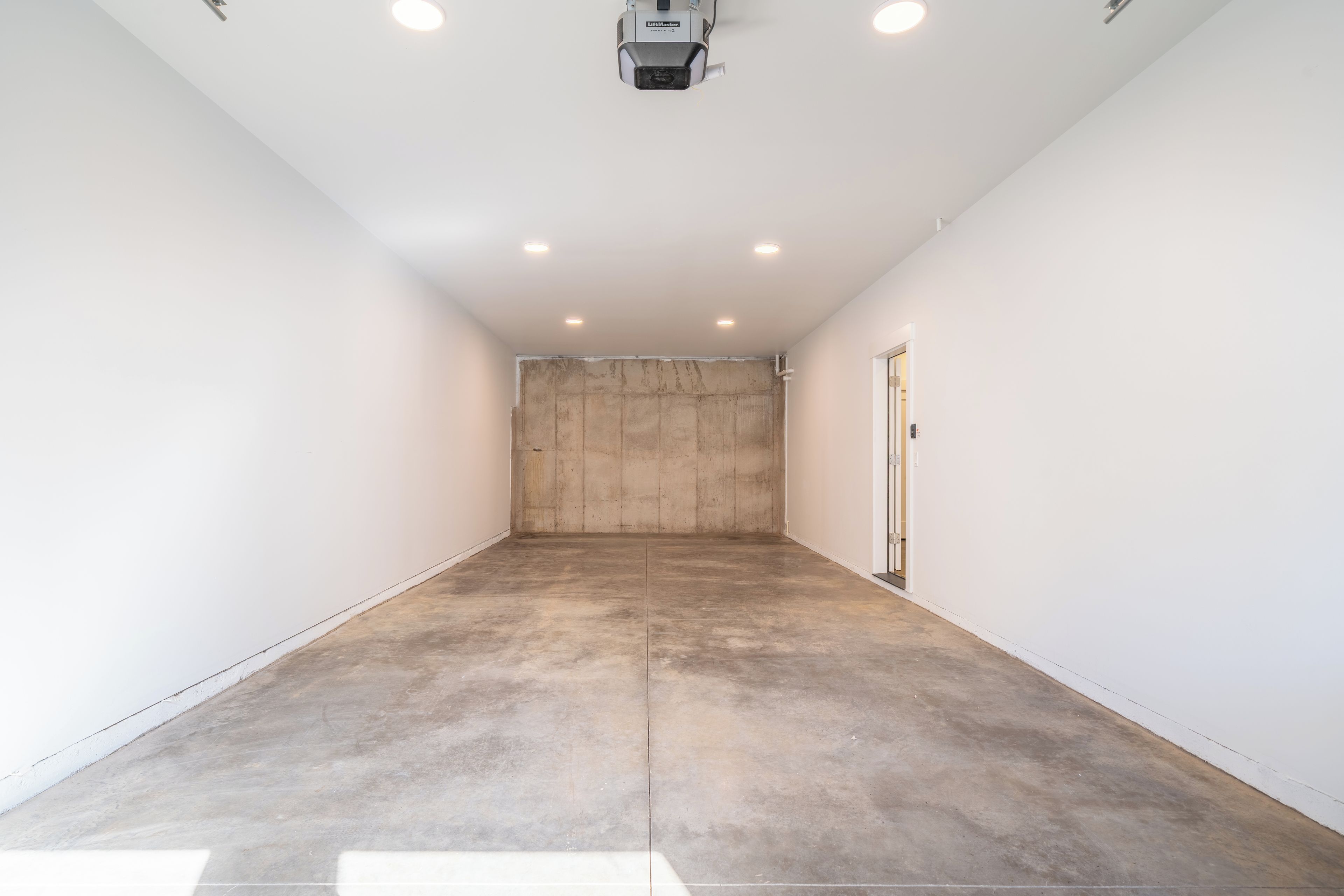 A spacious, empty room with white walls and a concrete floor, featuring recessed ceiling lights and a door on the right.