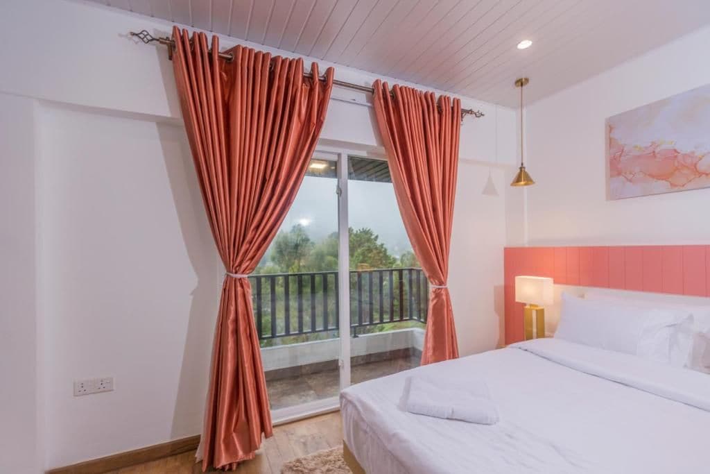 Habitación de hotel con cama, mesita de noche con lámpara, ventana con cortinas y vistas al exterior.