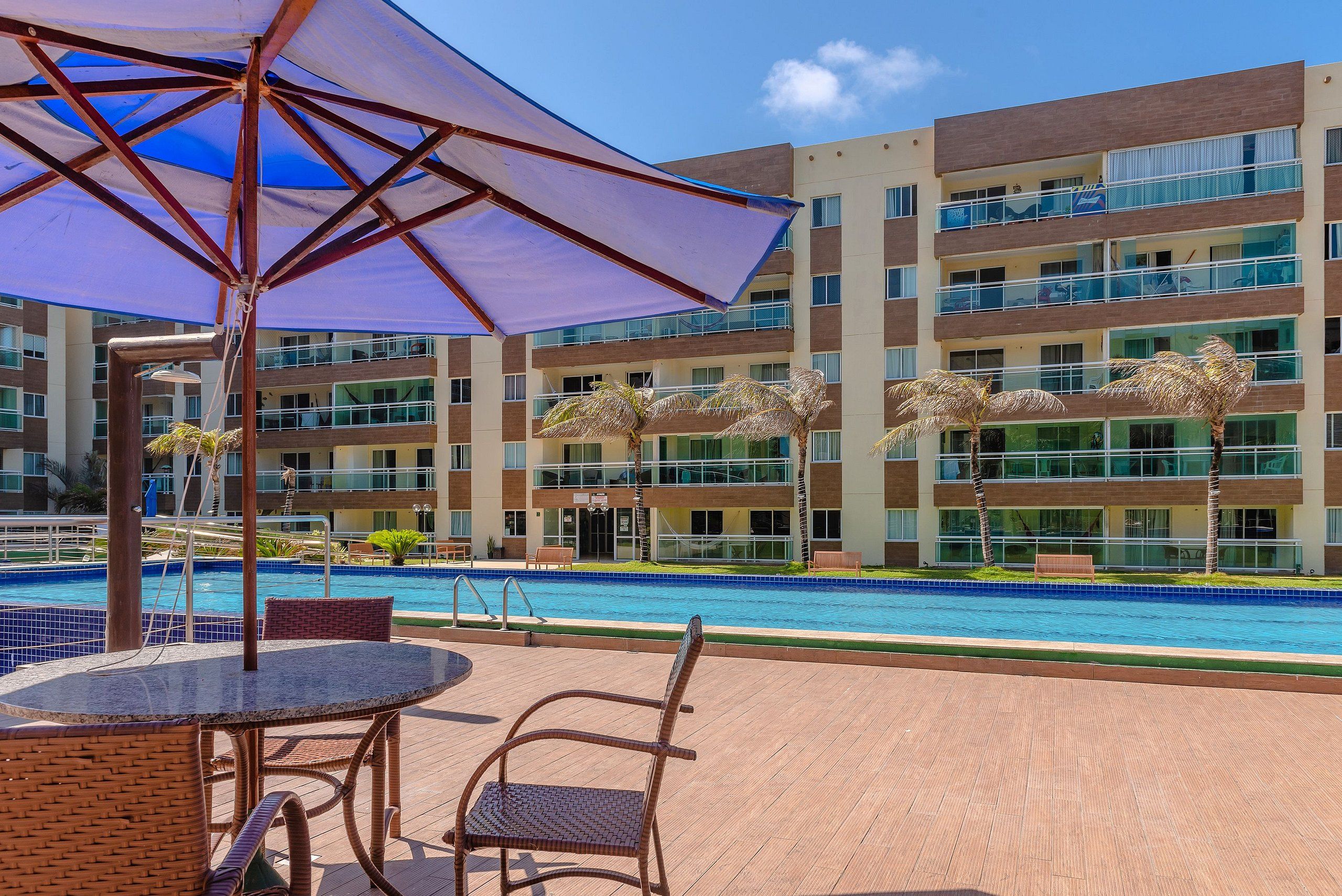 Zona junto a la piscina con una mesa y sillas, un parasol grande y un edificio con balcones al fondo.