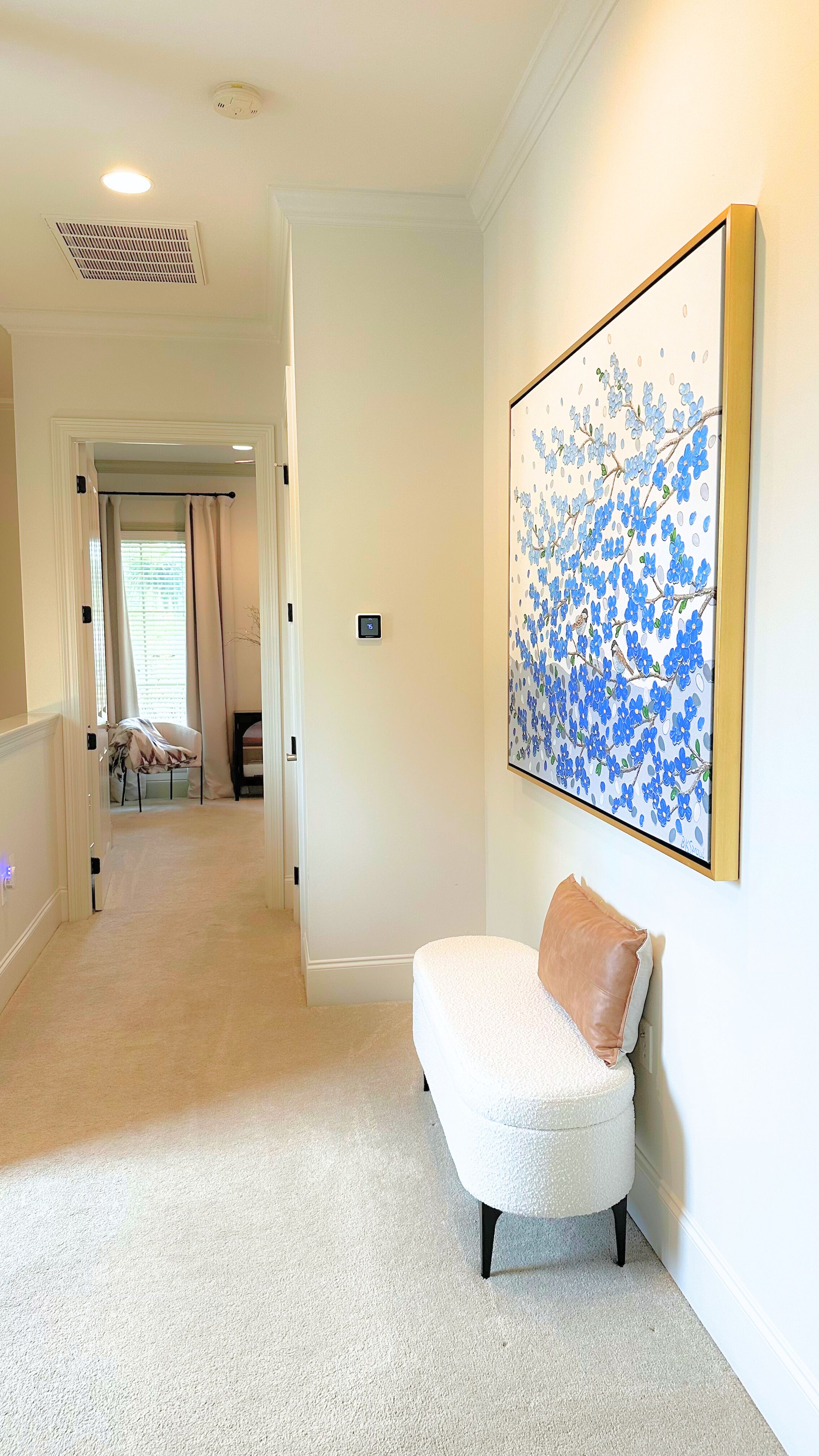 A hallway with a white chair, a large floral painting on the wall, and a door leading to another room.