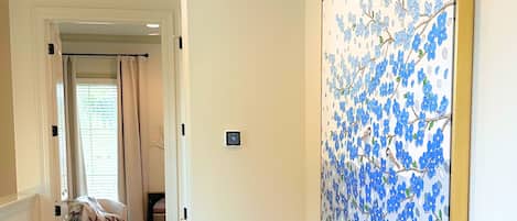 A hallway with a white chair, a large floral painting on the wall, and a door leading to another room.