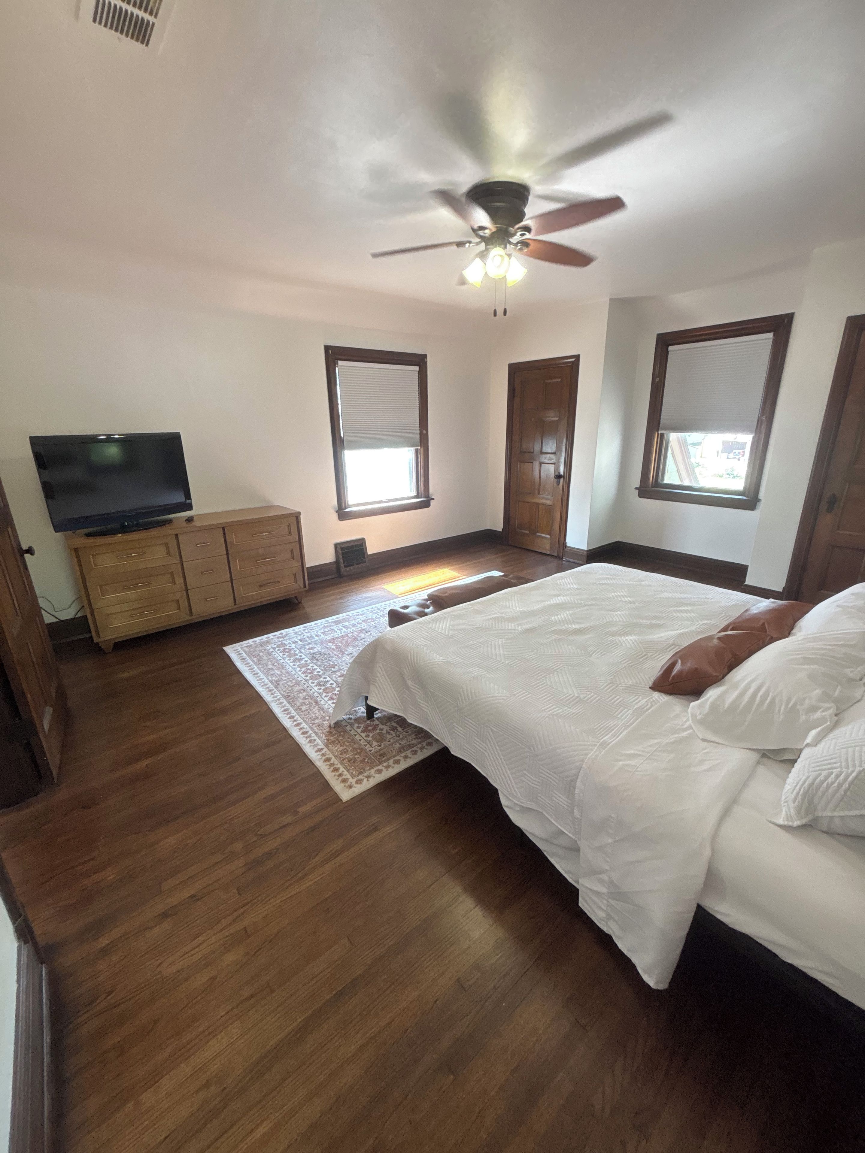 A bedroom with a bed, a television on a wooden cabinet, a ceiling fan, and a window with blinds.