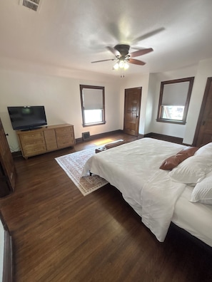 Une chambre à coucher avec un lit, un téléviseur posé sur un meuble en bois, un ventilateur de plafond et une fenêtre avec des stores.