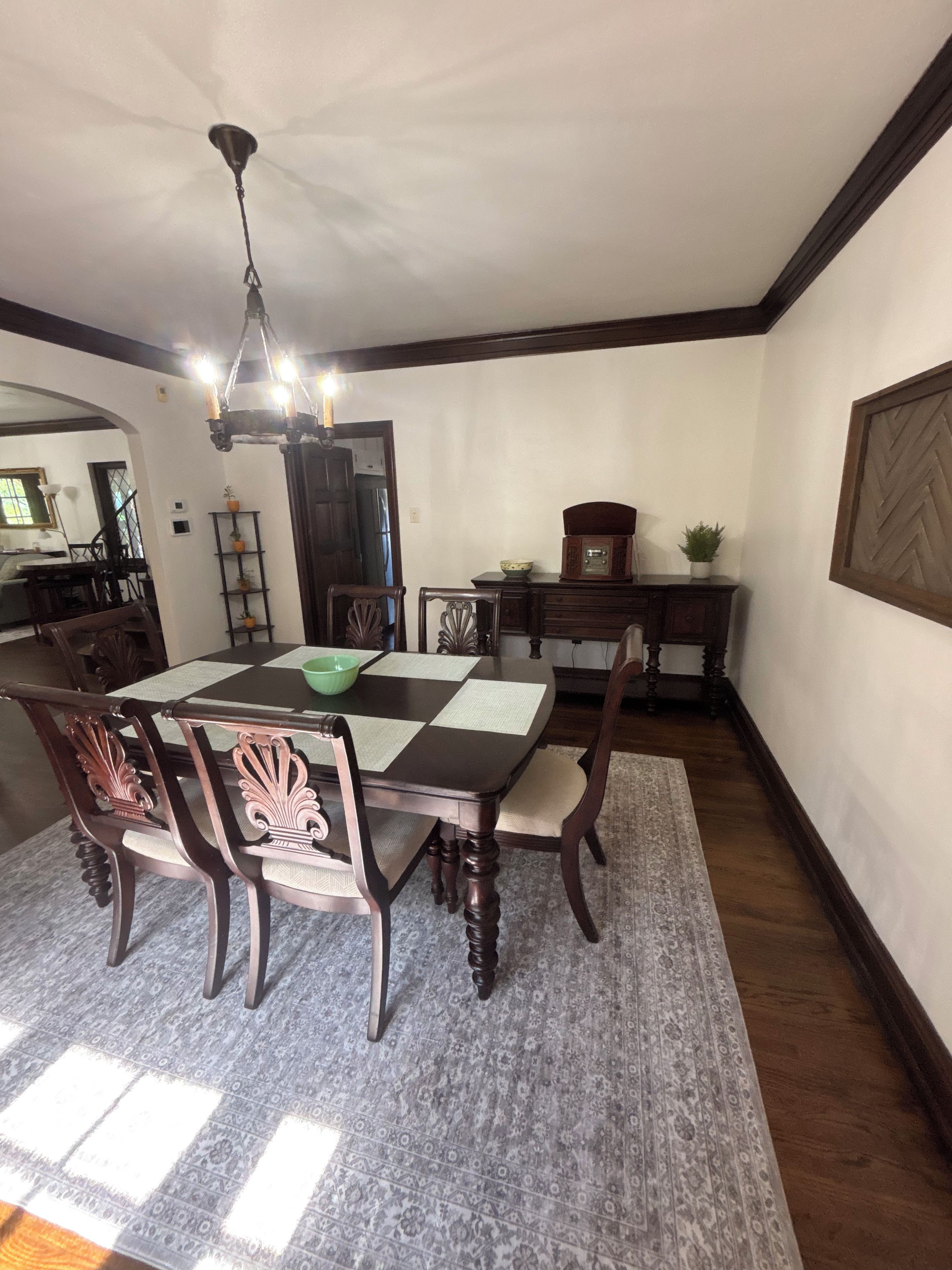 A dining area with a checkered table, wooden chairs, a chandelier, and a patterned rug.