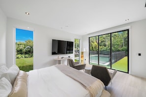 Ein modernes Hotelzimmer mit einem großen Bett, einem Schreibtisch, einem Sessel, einem Fernseher und Blick auf einen Swimmingpool und Garten.