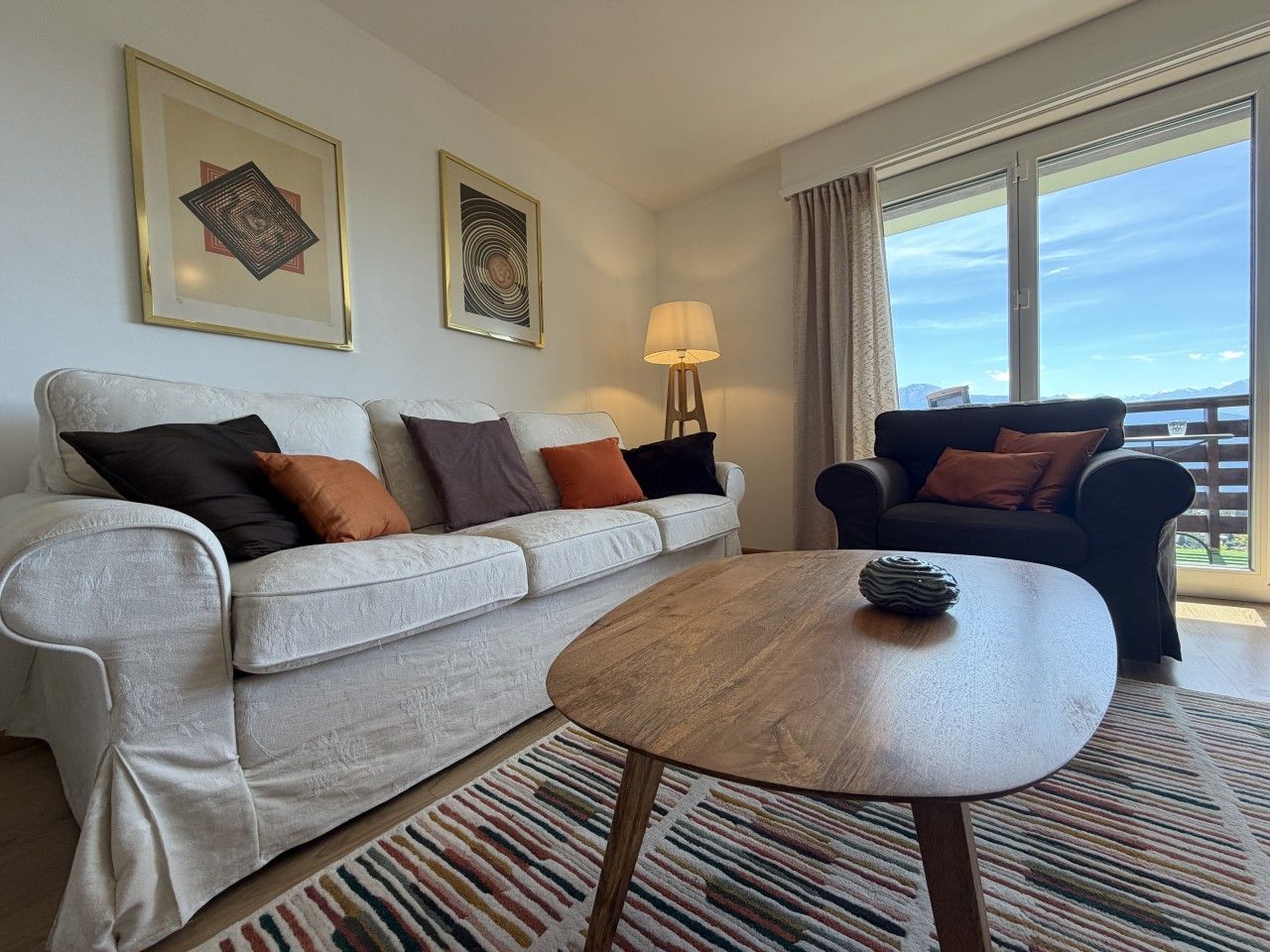 Una sala de estar moderna con un sofá, una mesa de centro y un sillón. Hay dos obras de arte enmarcadas en la pared, una lámpara y vistas al exterior a través de una ventana.