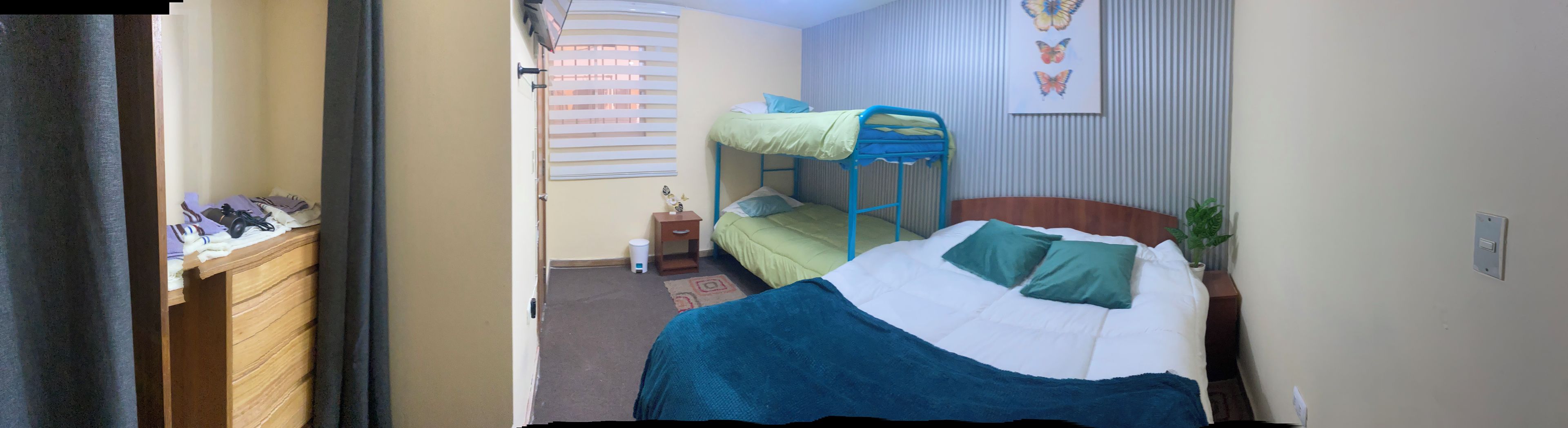 A dormitory room with bunk beds, a desk with personal items, a small bedside table, and a wall-mounted shelf.