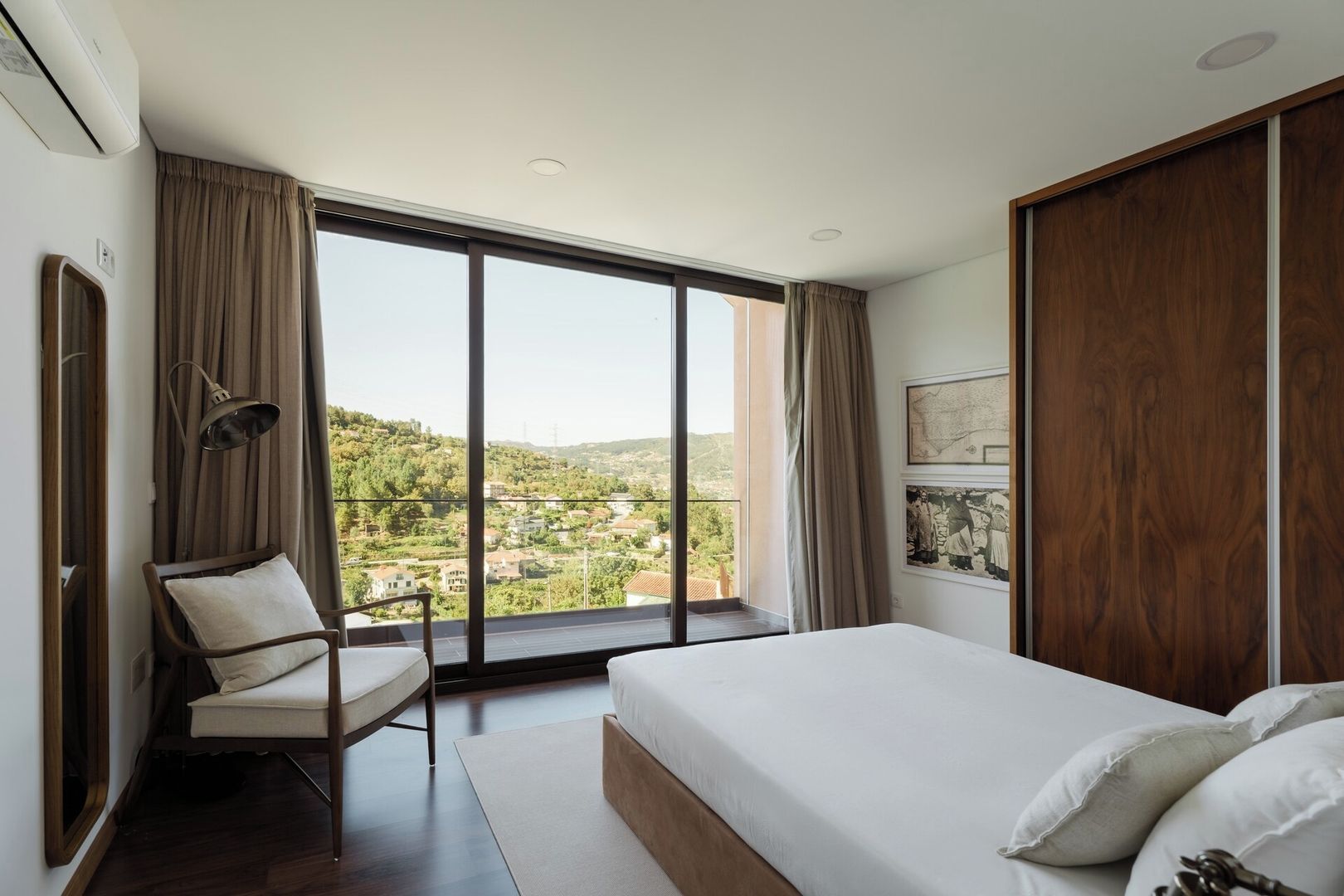 A bedroom with a large bed, a chair, a window with curtains, and a view of a town.