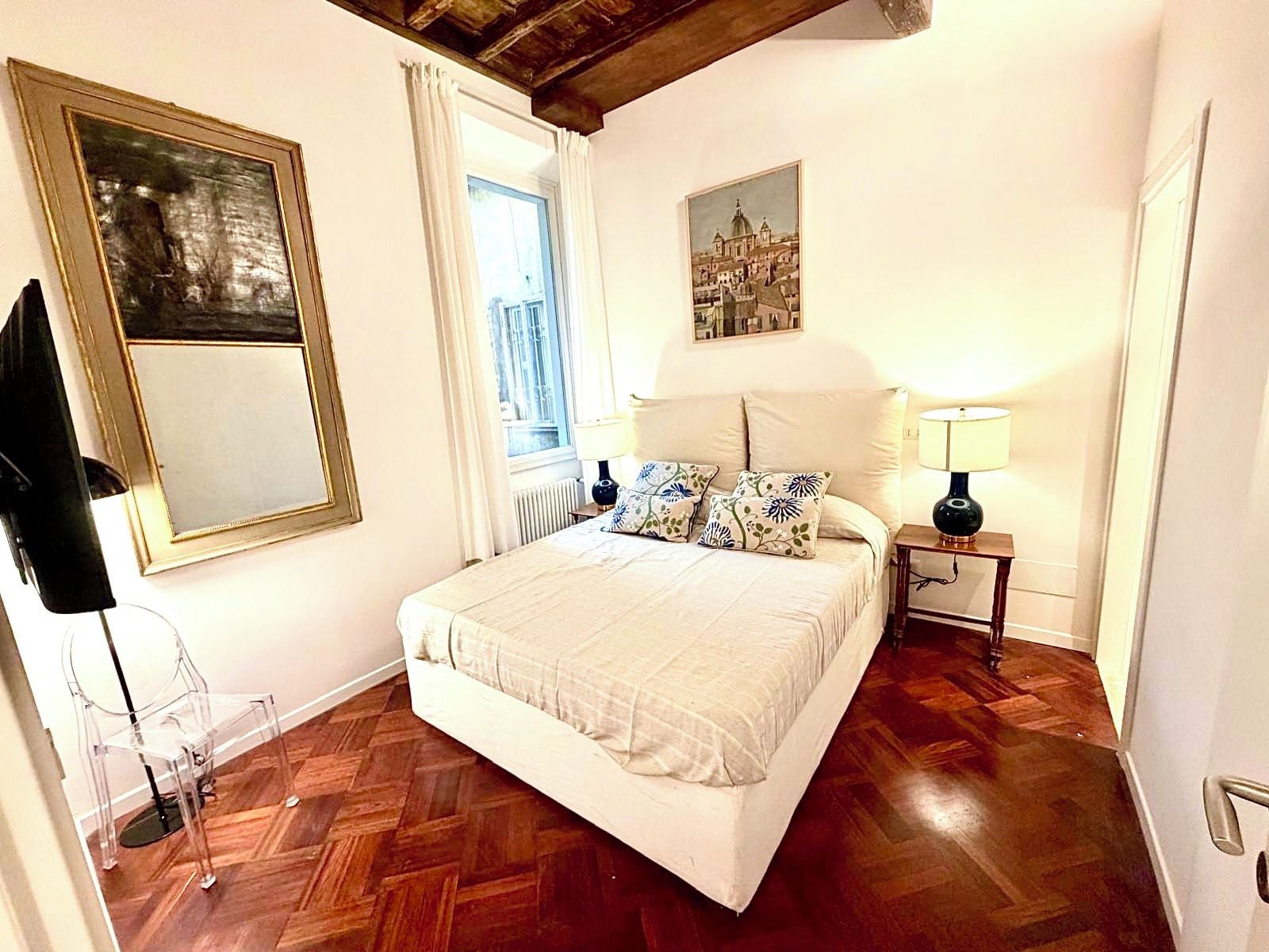 A bedroom with a bed, a nightstand, a lamp, a window with curtains, a mirror, and a picture on the wall.