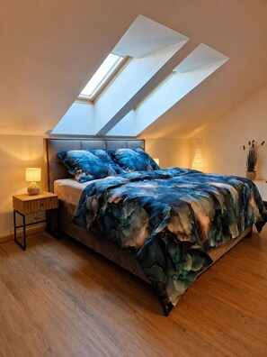 A bedroom with a sloped ceiling, a bed with a blue floral bedspread, a bedside table with a lamp, and a potted plant. - Loft Park Olomouc - Spacious 109 m² Loft with AC in City Centre (Olomouc)