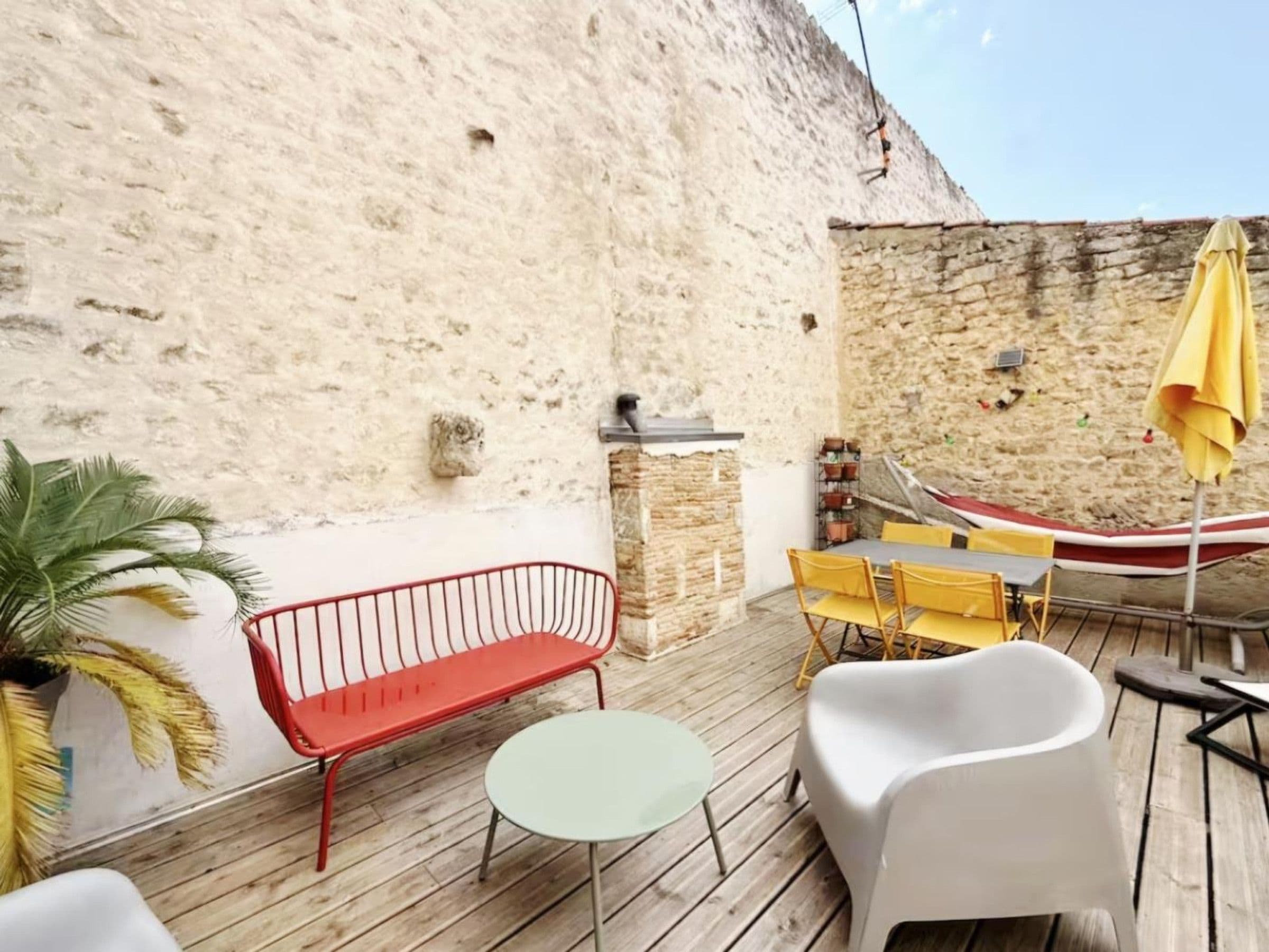 Un patio in legno con una panca rossa, una sedia bianca, un tavolino rotondo e un ombrellone giallo.