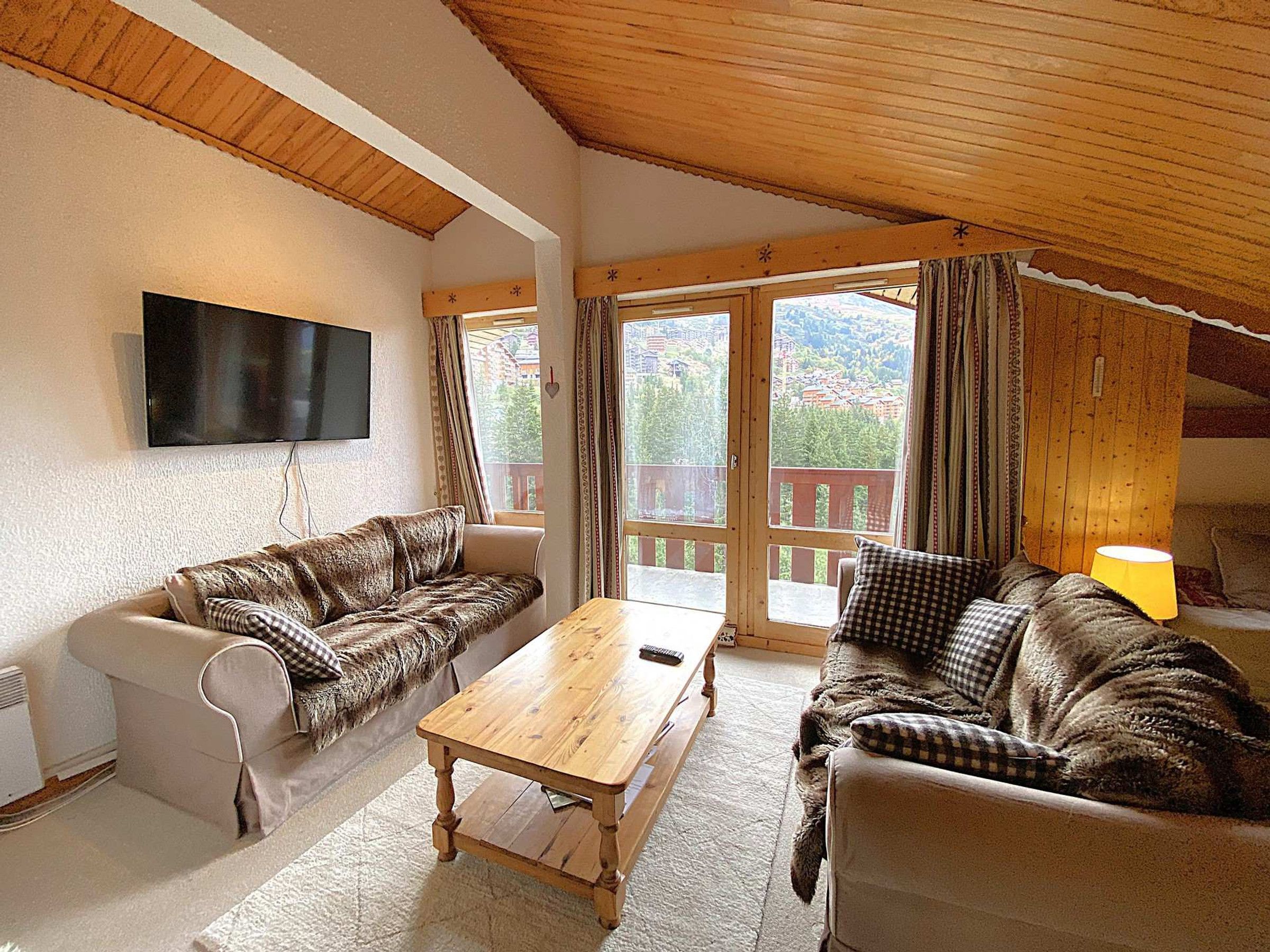 A cozy living room with a wooden ceiling, a flat-screen TV mounted on the wall, a wooden coffee table, and two sofas with checkered cushions.