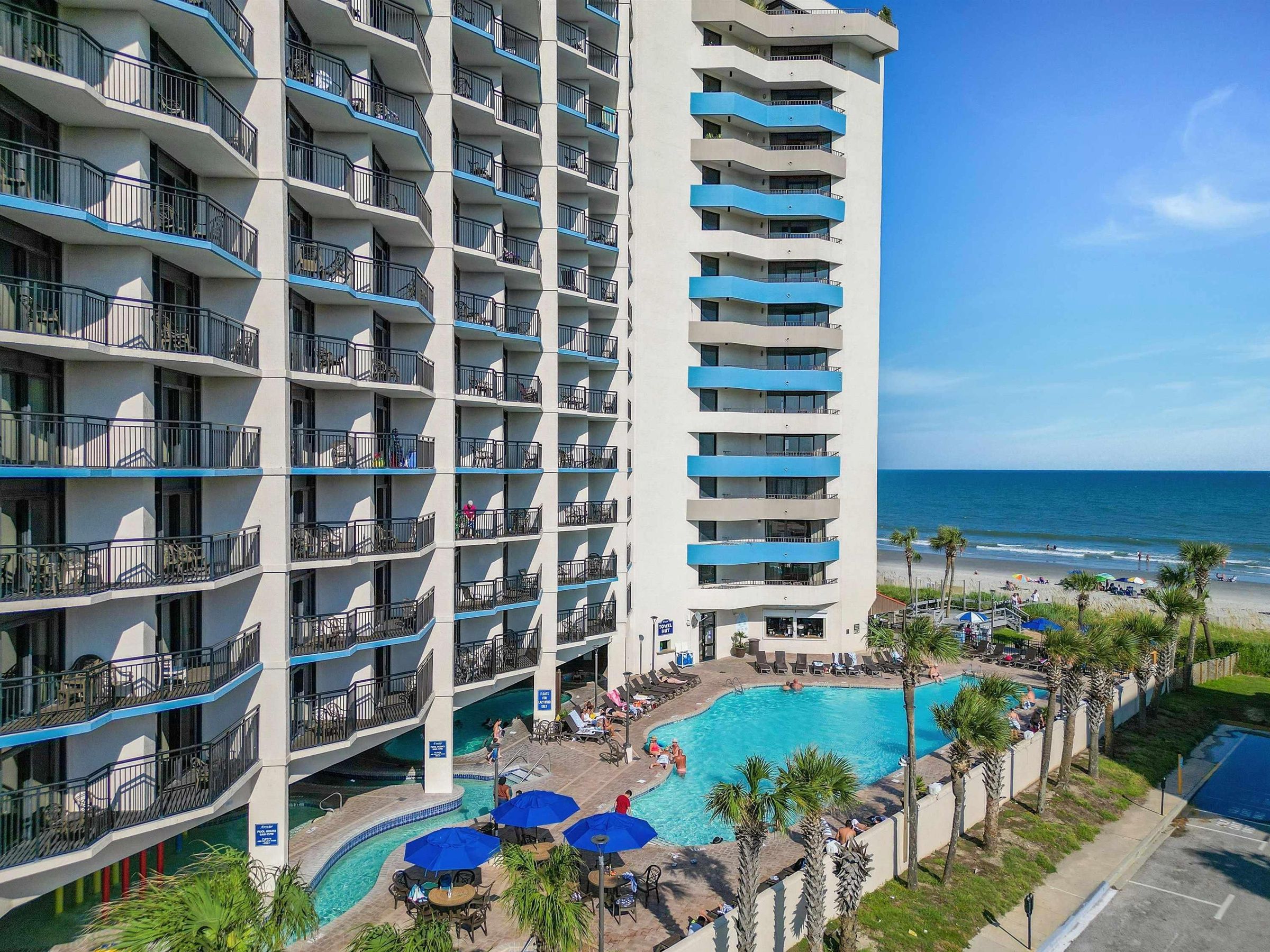 A multi-story hotel with a pool area, outdoor seating, and a view of the beach.