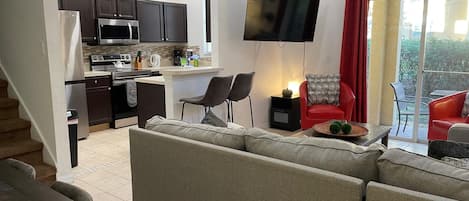 A modern living room with a sofa, a dining table, and a kitchen area with dark cabinets and a flat-screen TV.