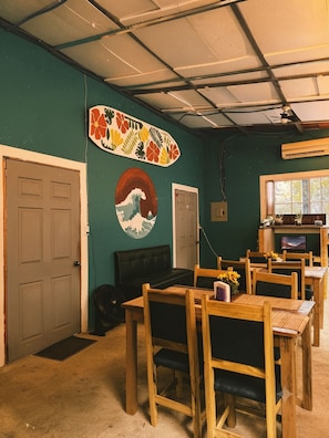 Une pièce décorée avec une œuvre d’art représentant une planche de surf, un tableau représentant une vague, et un coin repas avec des tables et des chaises en bois.