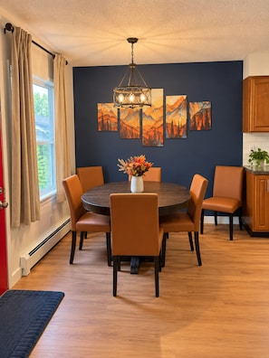 Un comedor con una mesa redonda, seis sillas, un jarrón con flores y una pared decorada con tres cuadros.