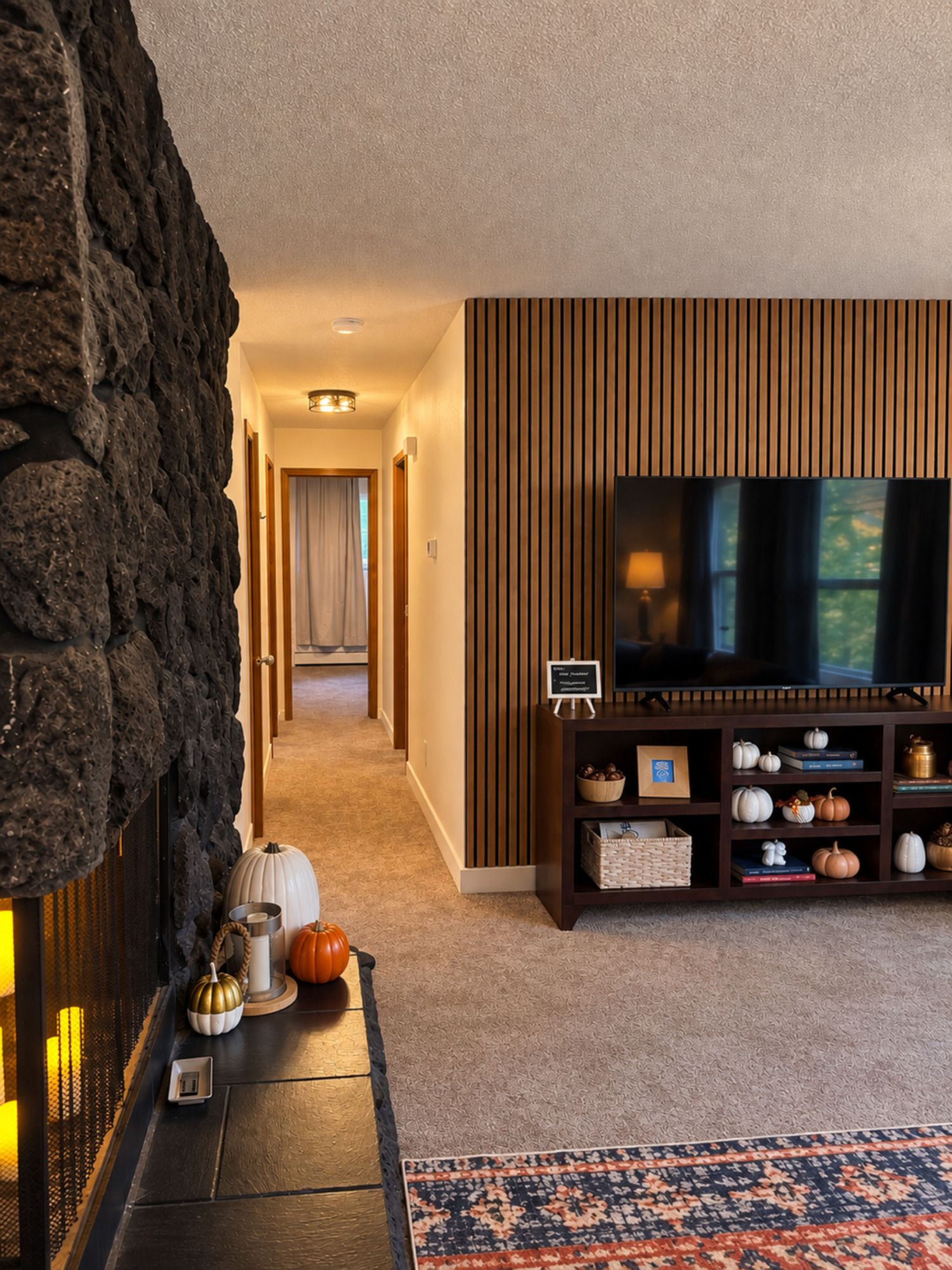 Een moderne woonkamer met een stenen openhaard, een flatscreen televisie op een houten standaard, en een decoratieve plank met pompoenen.