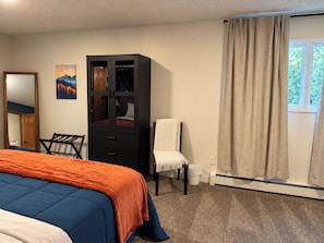Un dormitorio con cama, un armario, una silla, una ventana con cortinas y un cuadro en la pared.