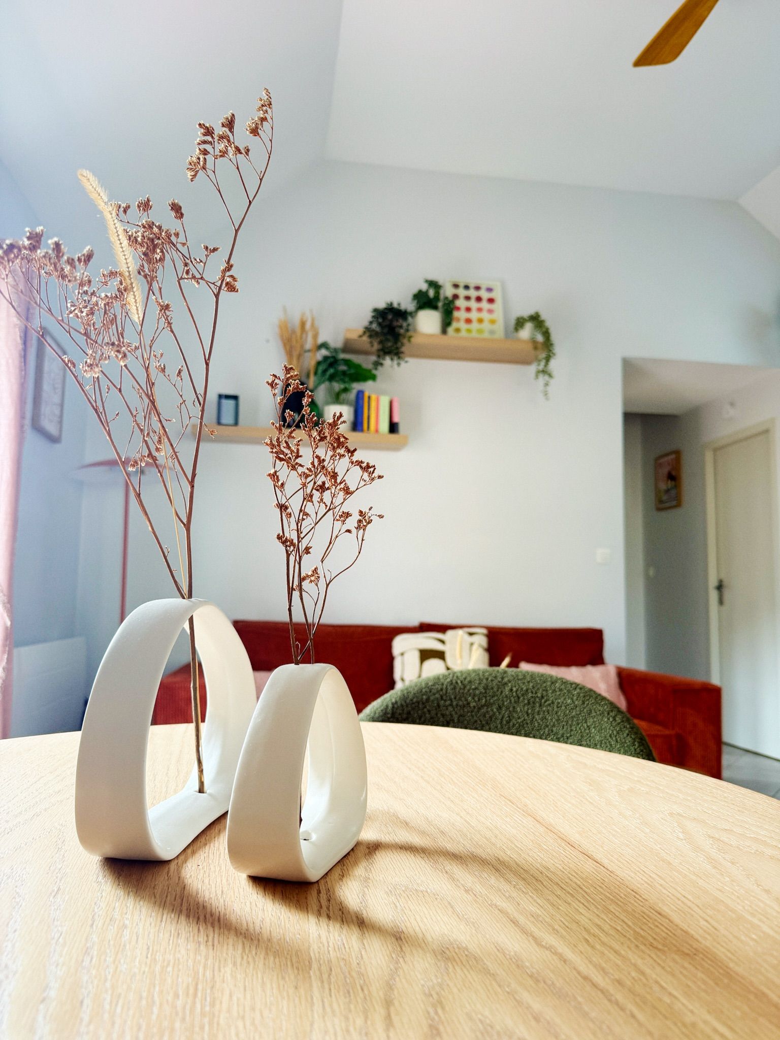 Une pièce avec une table en bois, un vase blanc contenant des fleurs séchées, un canapé rouge et une bibliothèque en arrière-plan.