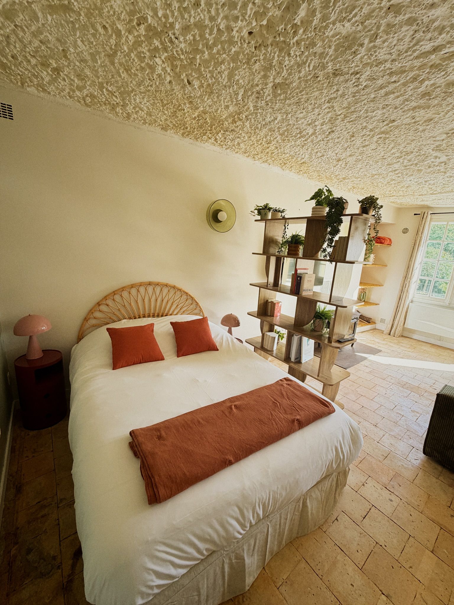 A bedroom with a bed, a bedside table, a lamp, a shelf with plants, and a window.