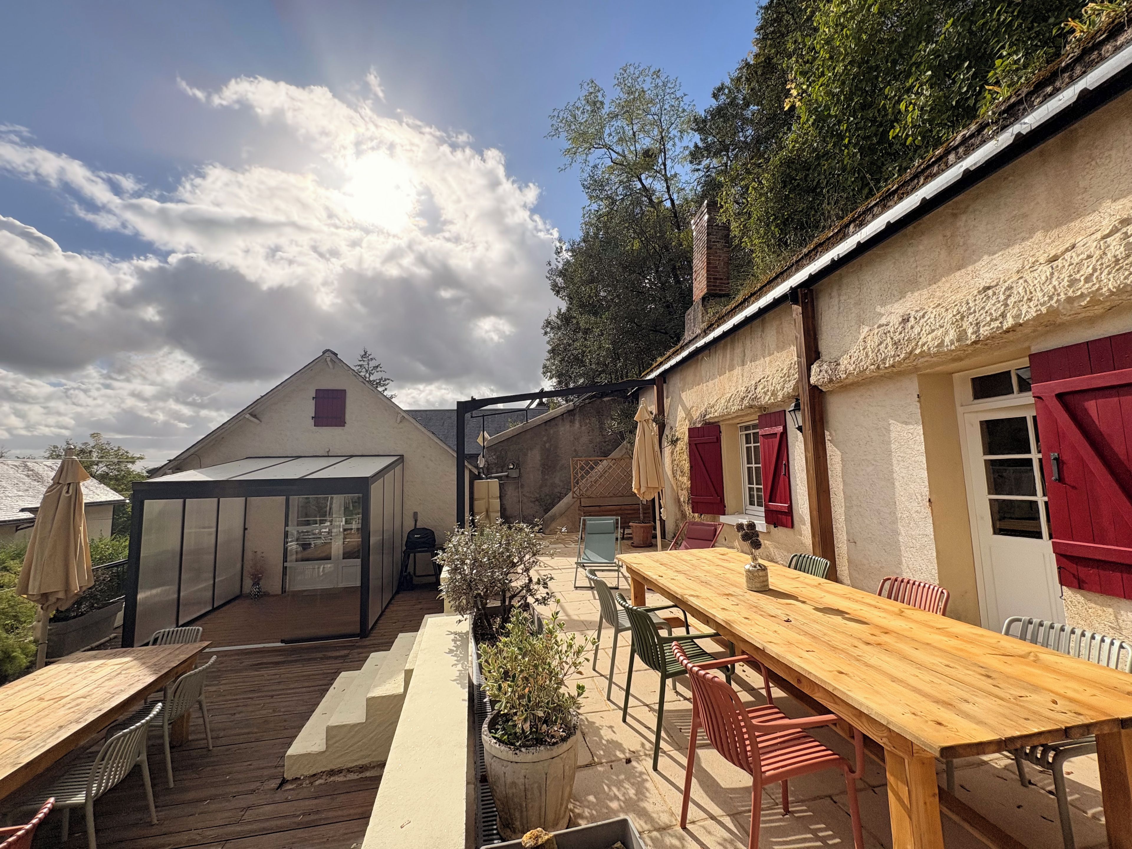 A patio with a wooden table and chairs, a greenhouse, and a building with red shutters.